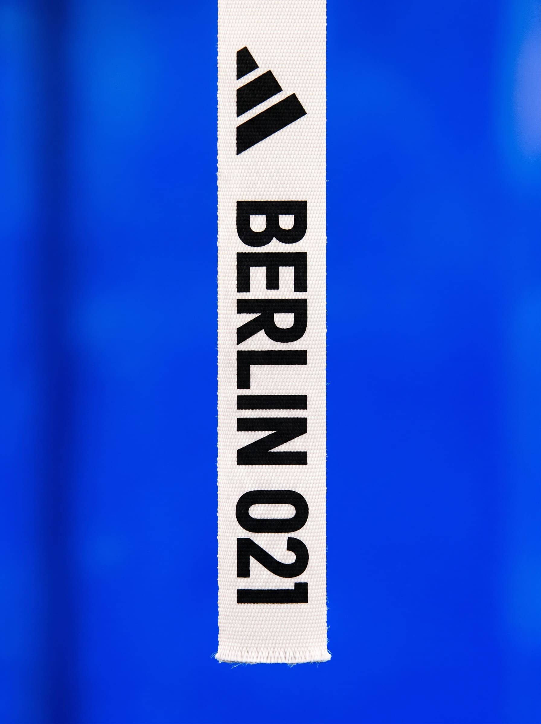 White woven strap printed with a stepped three-bar wedge mark, BERLIN, and 021 in black, hanging against a blurred blue field