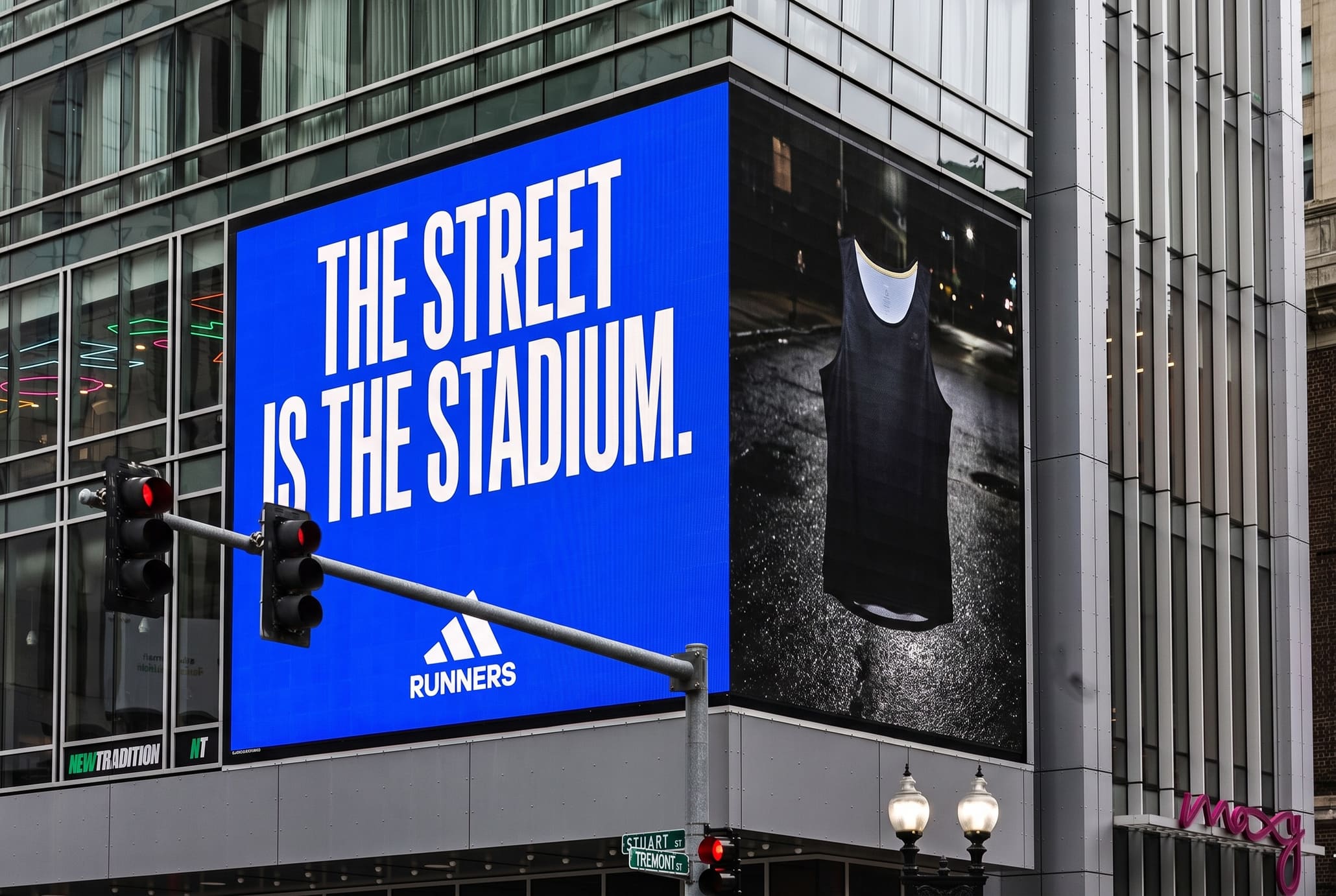 Corner LED billboard on a city building, blue face reading THE STREET IS THE STADIUM beside a right face showing a ghost-mannequin running singlet lit at night on wet black asphalt