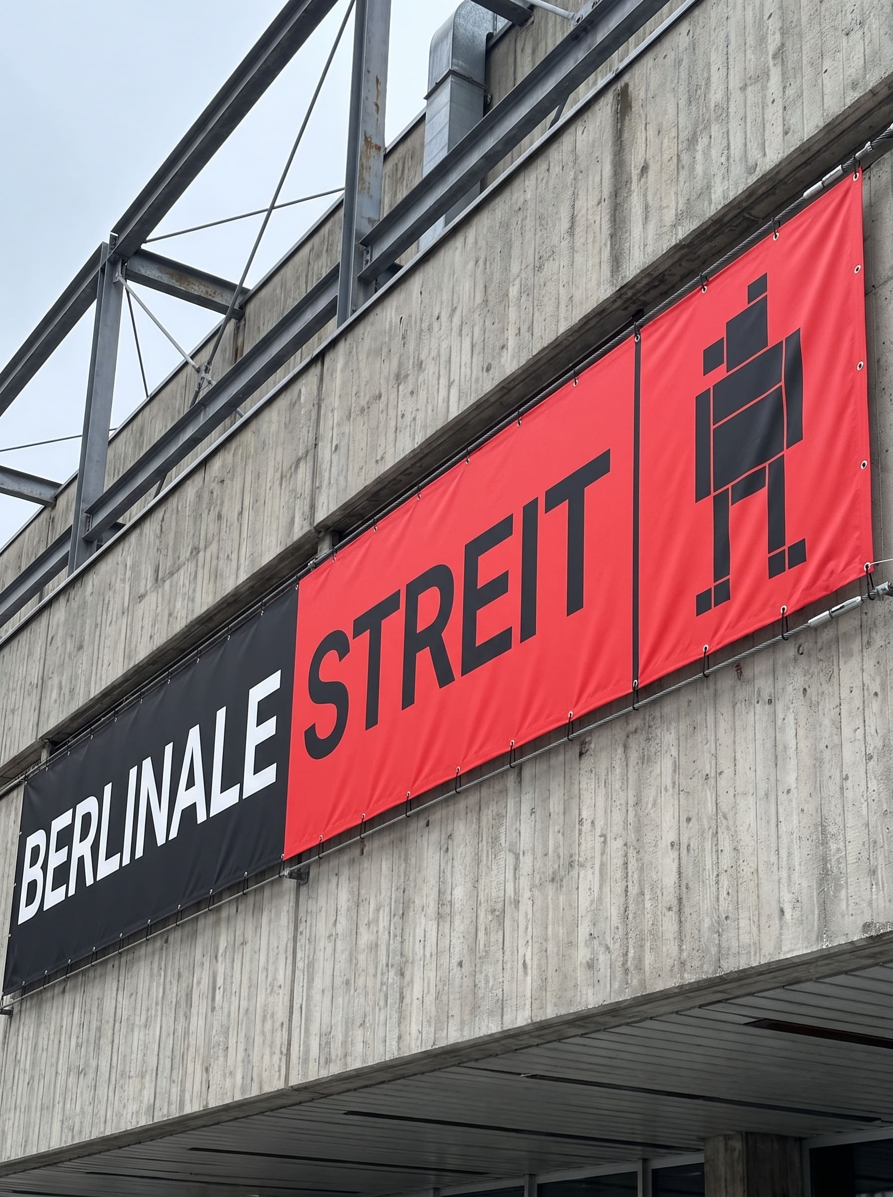 Red and black banner hung taut on steel cable along a concrete cinema facade reading BERLINALE STREIT, with the bear shown as a hand-cut silhouette rearing on its hind legs, forelimbs raised, in profile