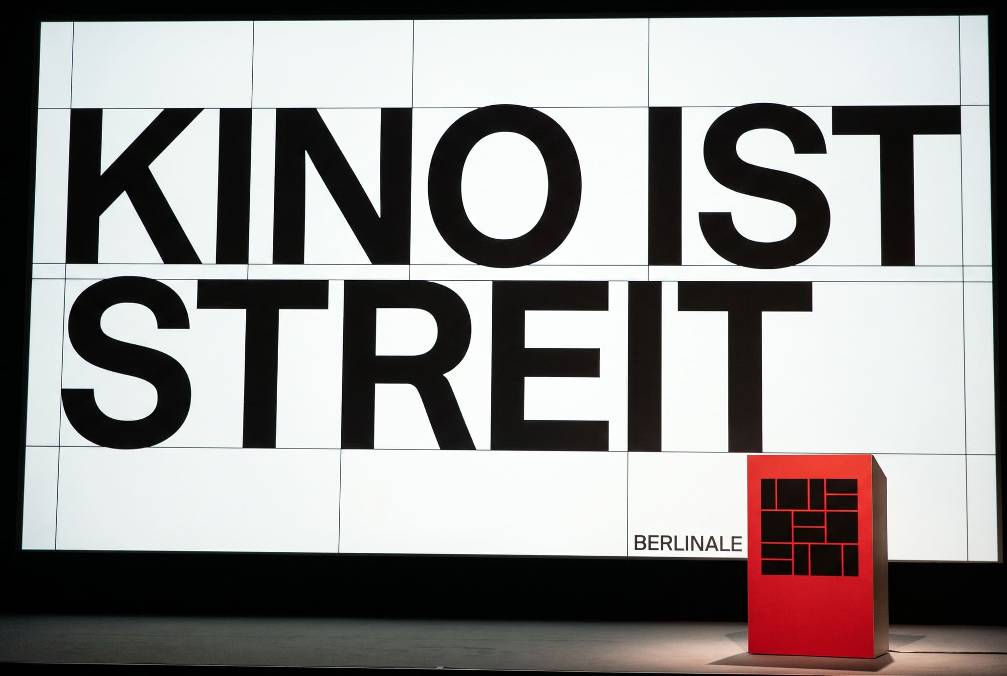 A red lectern bearing the geometric bear mark stands alone before a screen reading KINO IST STREIT