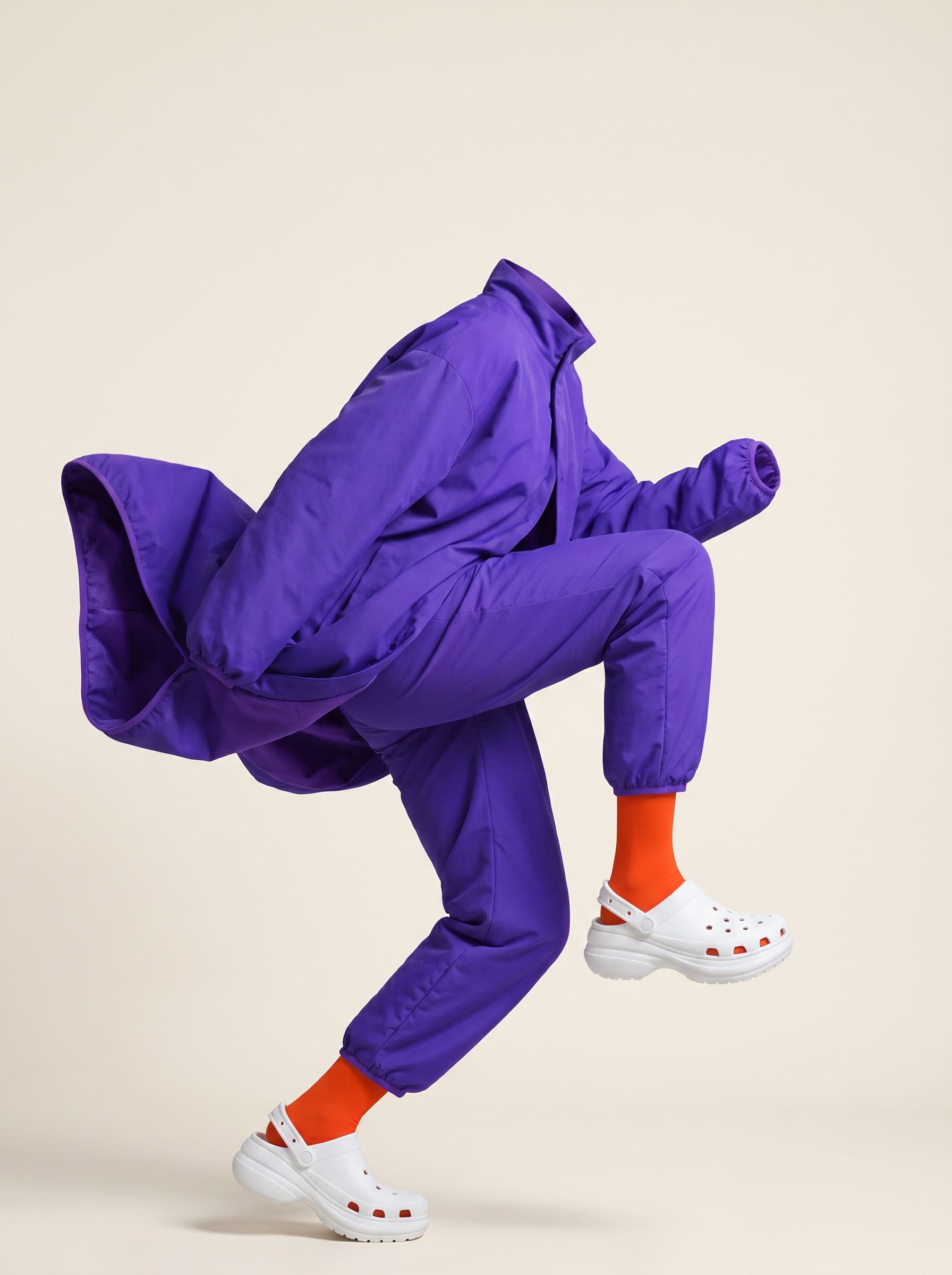 A hollow ghost-mannequin coverall in ultraviolet with orange tights and white moulded clogs, posed mid-stride and standing upright on a cream seamless floor, its own construction and footwear bearing the full weight of the form.