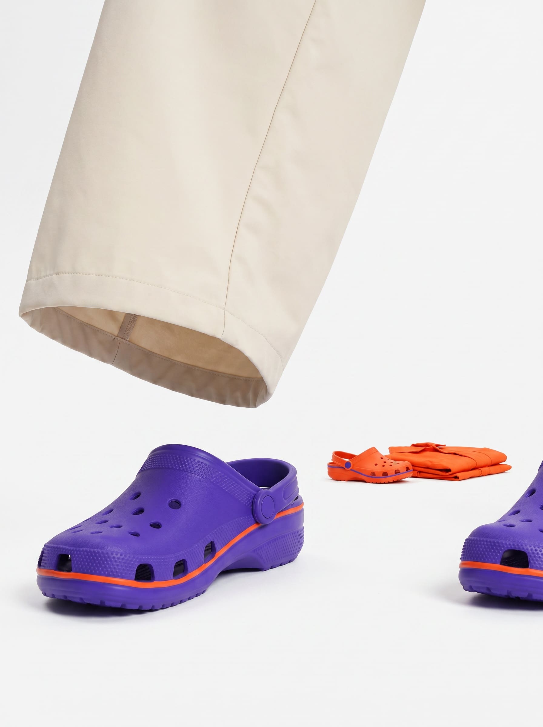 Giant cream ghost-mannequin trouser leg and ultraviolet clog resting on the studio floor in the foreground, with a small orange clog and folded coverall set on a plinth behind.