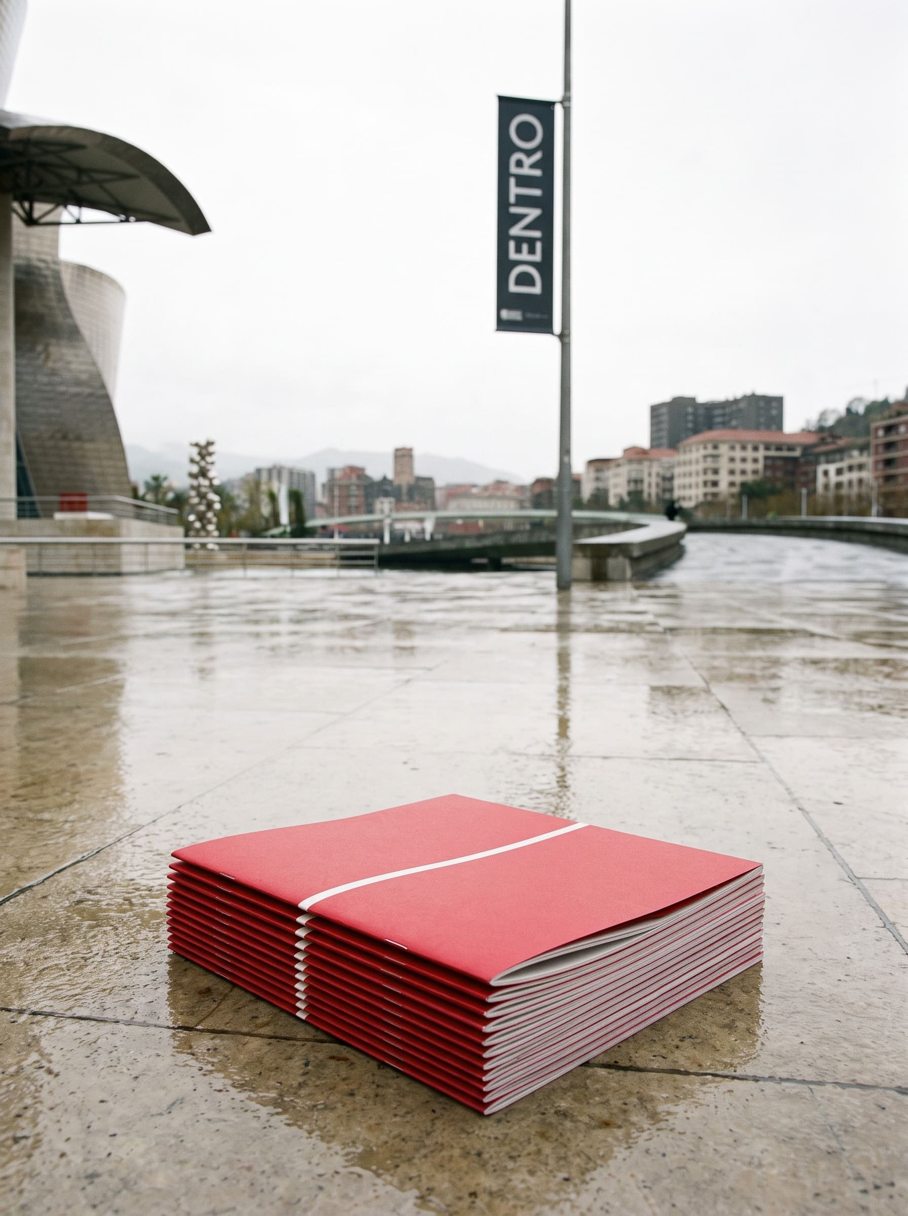 Overcast riverside plaza with a stack of folded red programmes resting flat on wet paving stones, museum cropped to a sliver