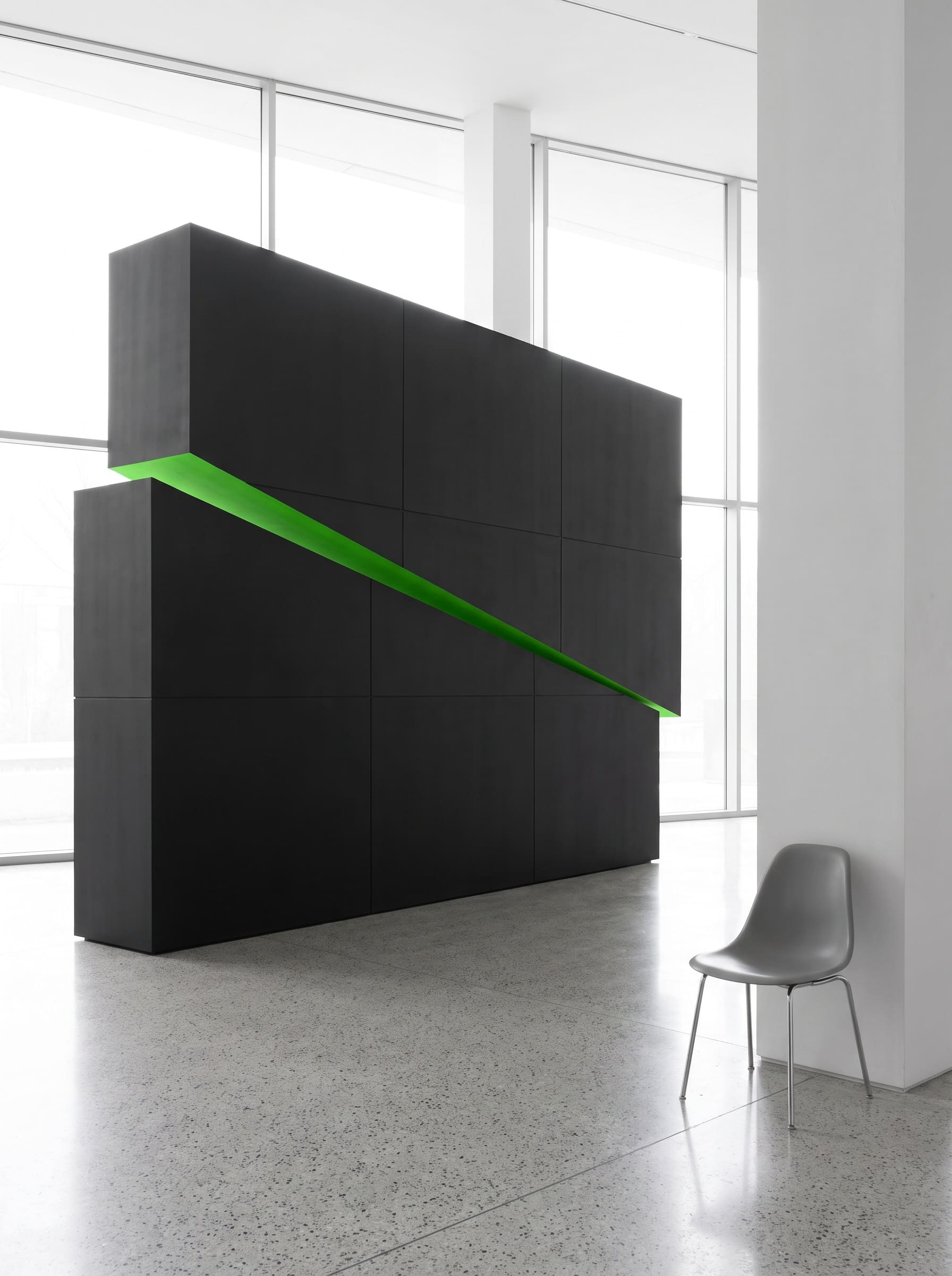Matte black wedge sculpture with an acid green cut face standing on terrazzo in a bright glass-walled atrium, an empty grey chair beside it for scale.