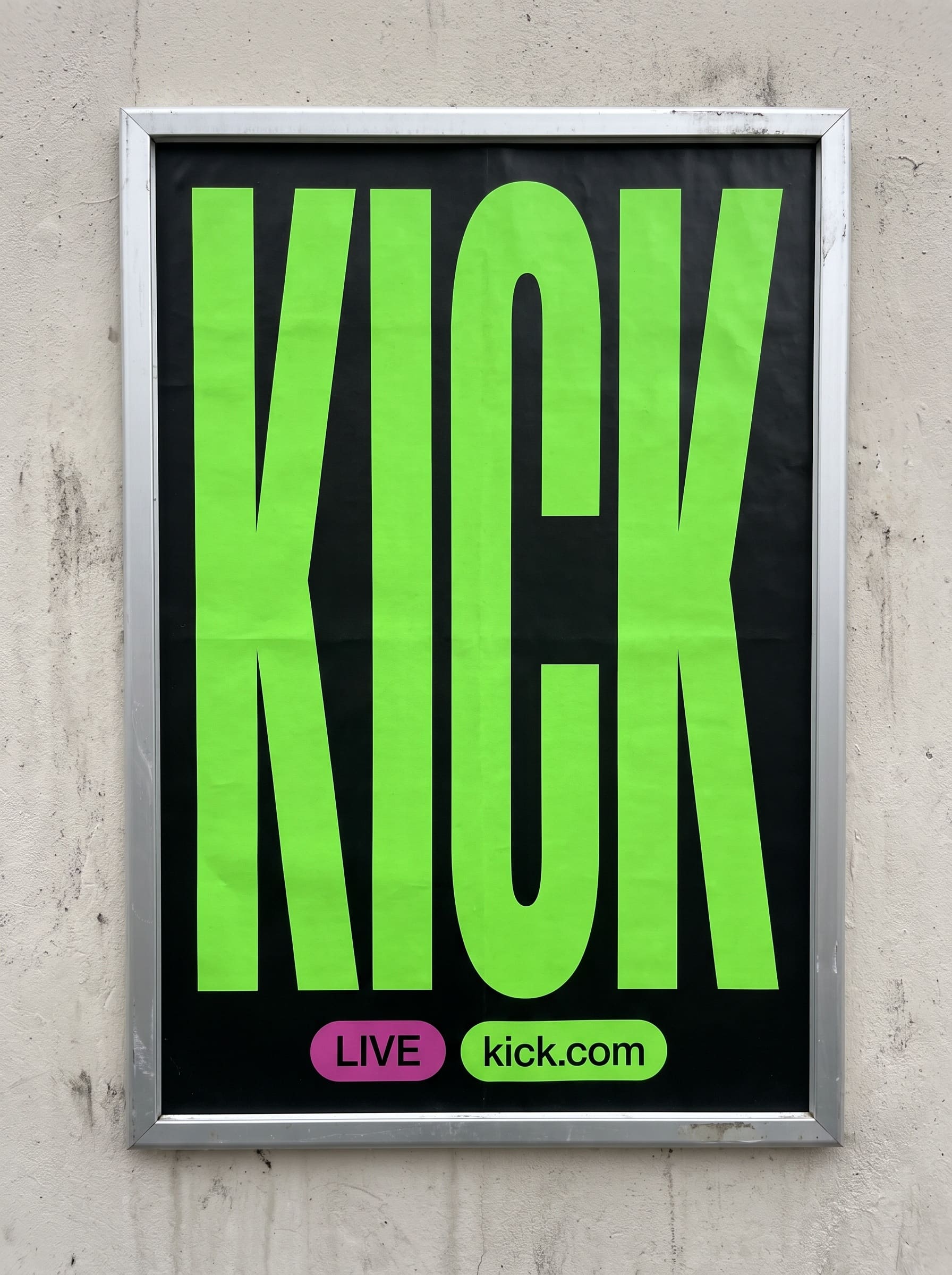 Framed poster on a pale wall: a green KICK wordmark stretched full height on black, two pills below.