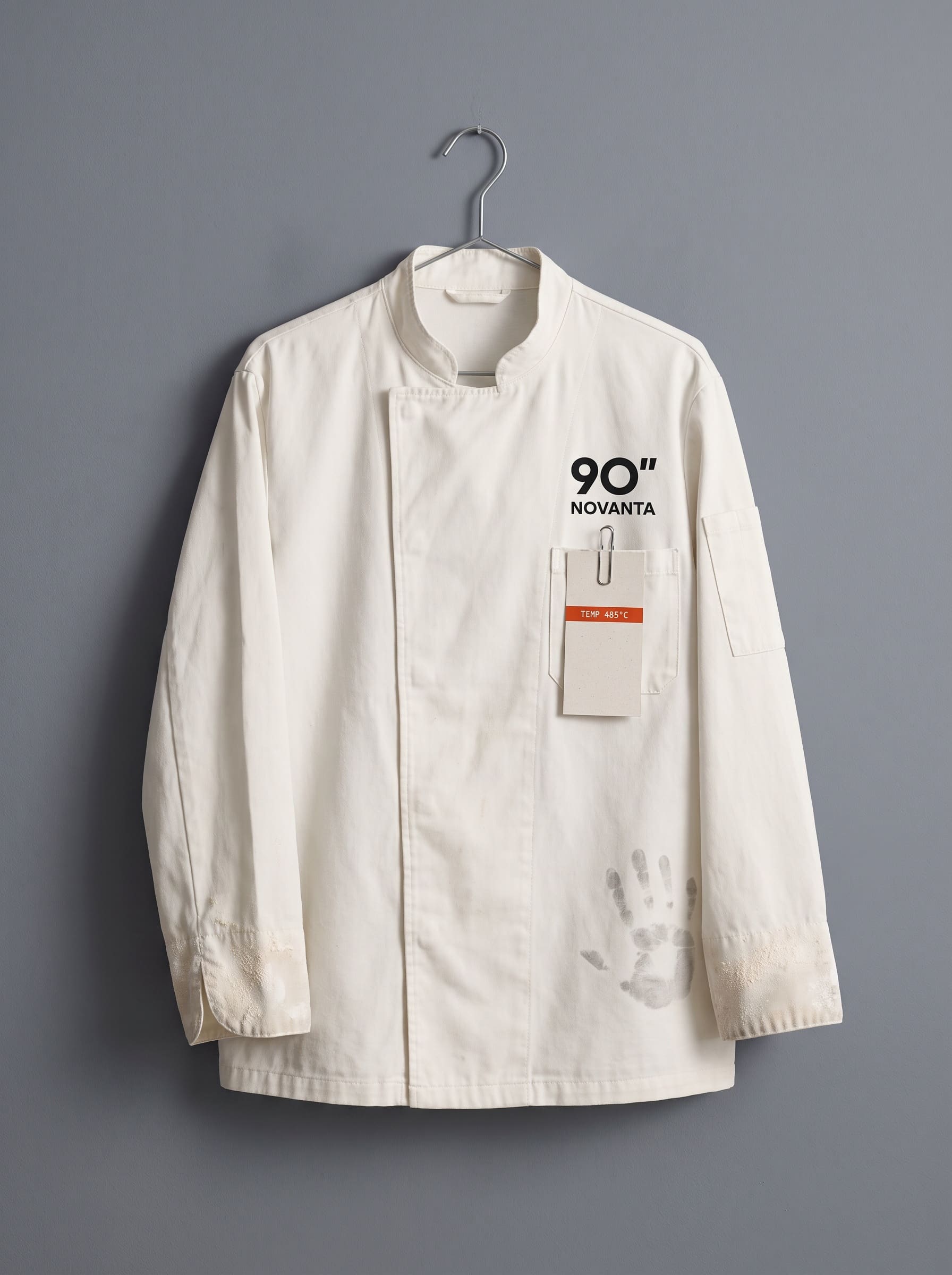 A white cook jacket hung on a steel hook against a grey wall, a docket clipped to its pocket