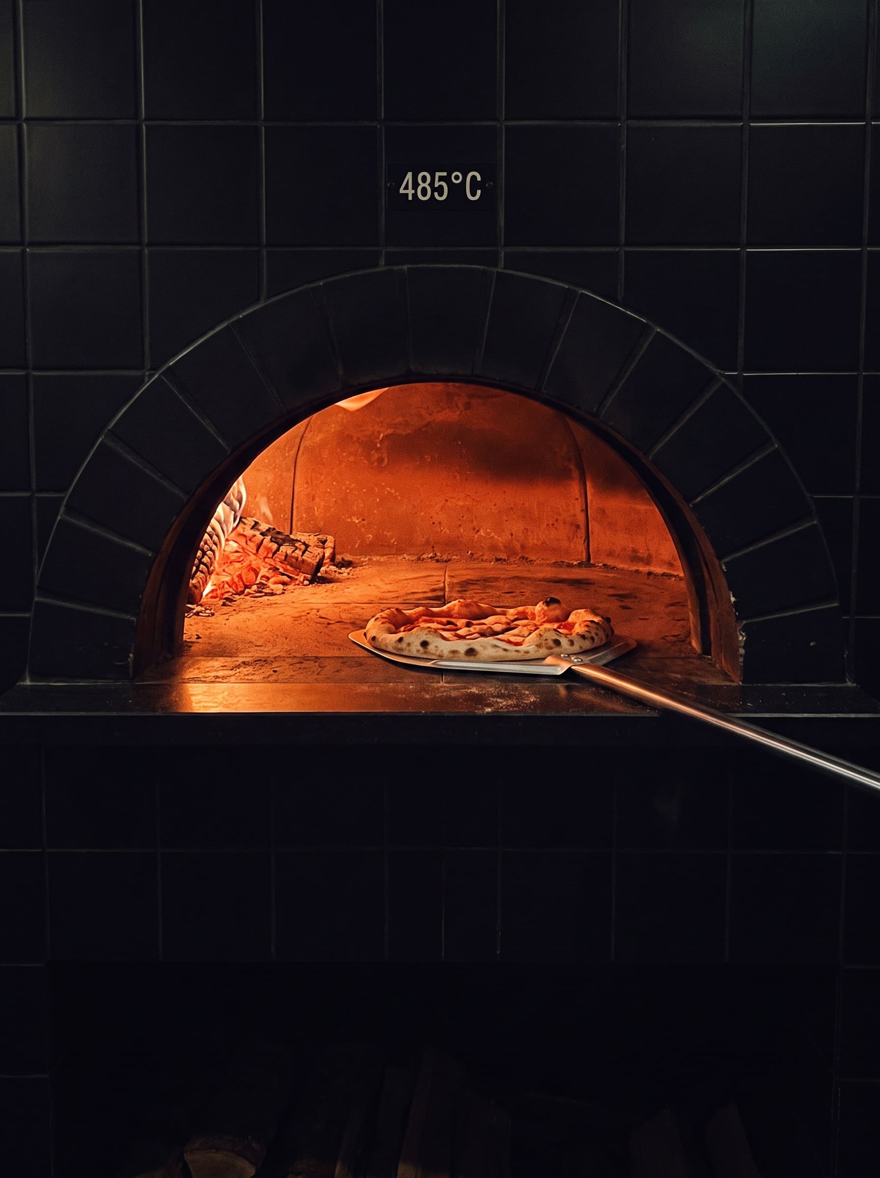The mouth of a domed pizza oven glowing orange, a steel peel sliding a pizza inside
