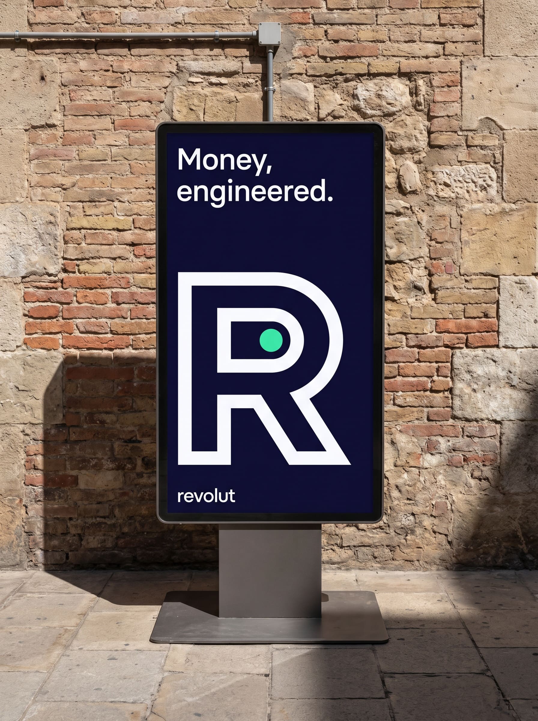 A freestanding double-sided digital totem stands self-supported on its weighted base plate on stone paving in a passage, its straight-on screen displaying an indigo poster reading Money, engineered above a large single-stroke R monogram and the revolut wordmark, its shadow cast across the paving to its left
