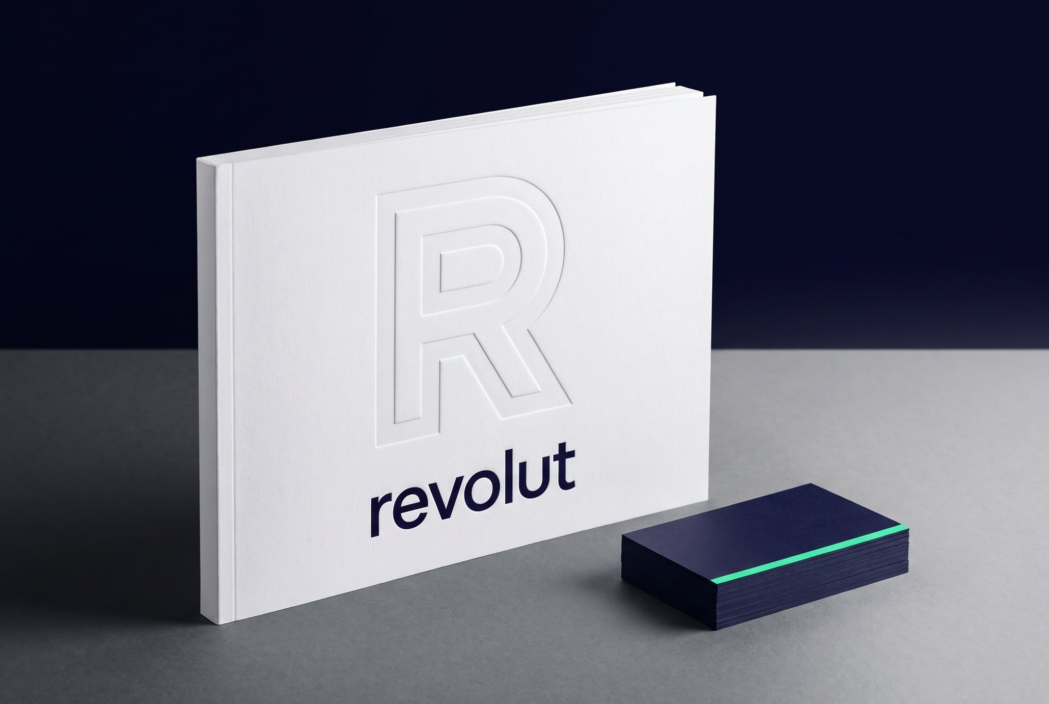 A cold white brand manual standing upright with a blind-debossed R on its cover, beside a small stack of indigo business cards topped with a mint edge-painted card, on a reflective surface
