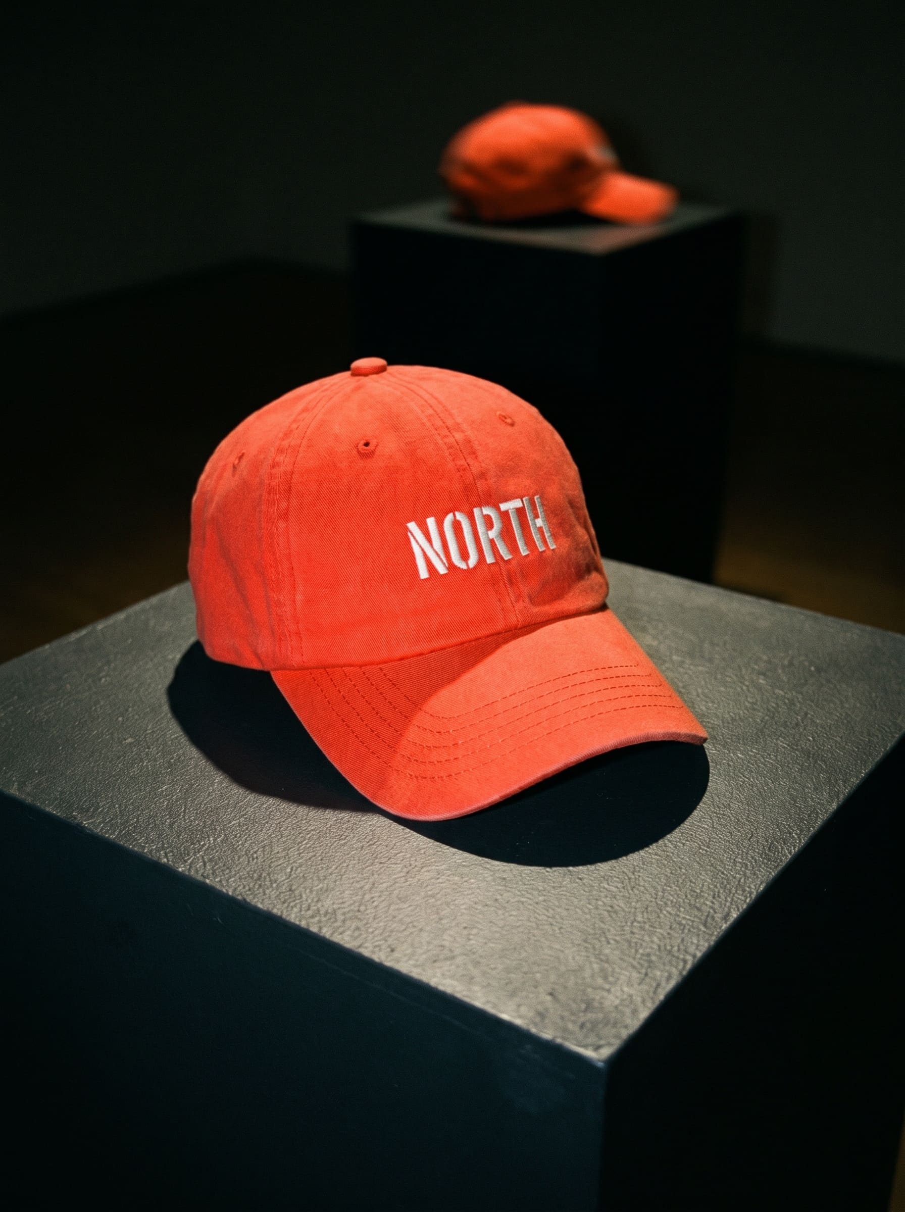 Flash-lit photograph of a red cap standing on a plinth, embroidered with a small NORTH, with a second cap blurred on a plinth behind it