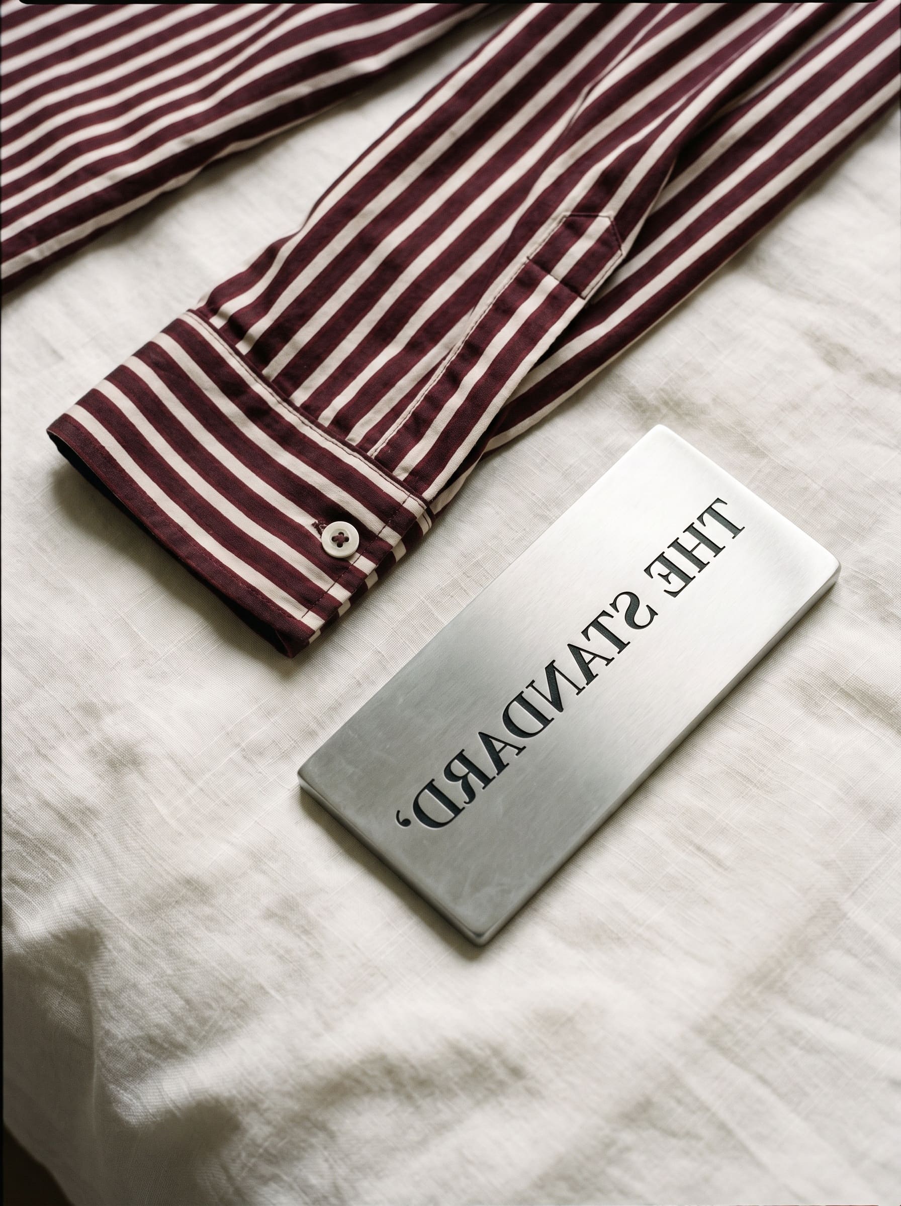 A folded oxblood-and-cream striped pyjama cuff resting on cream linen beside a chrome key fob engraved with an upside-down wordmark.