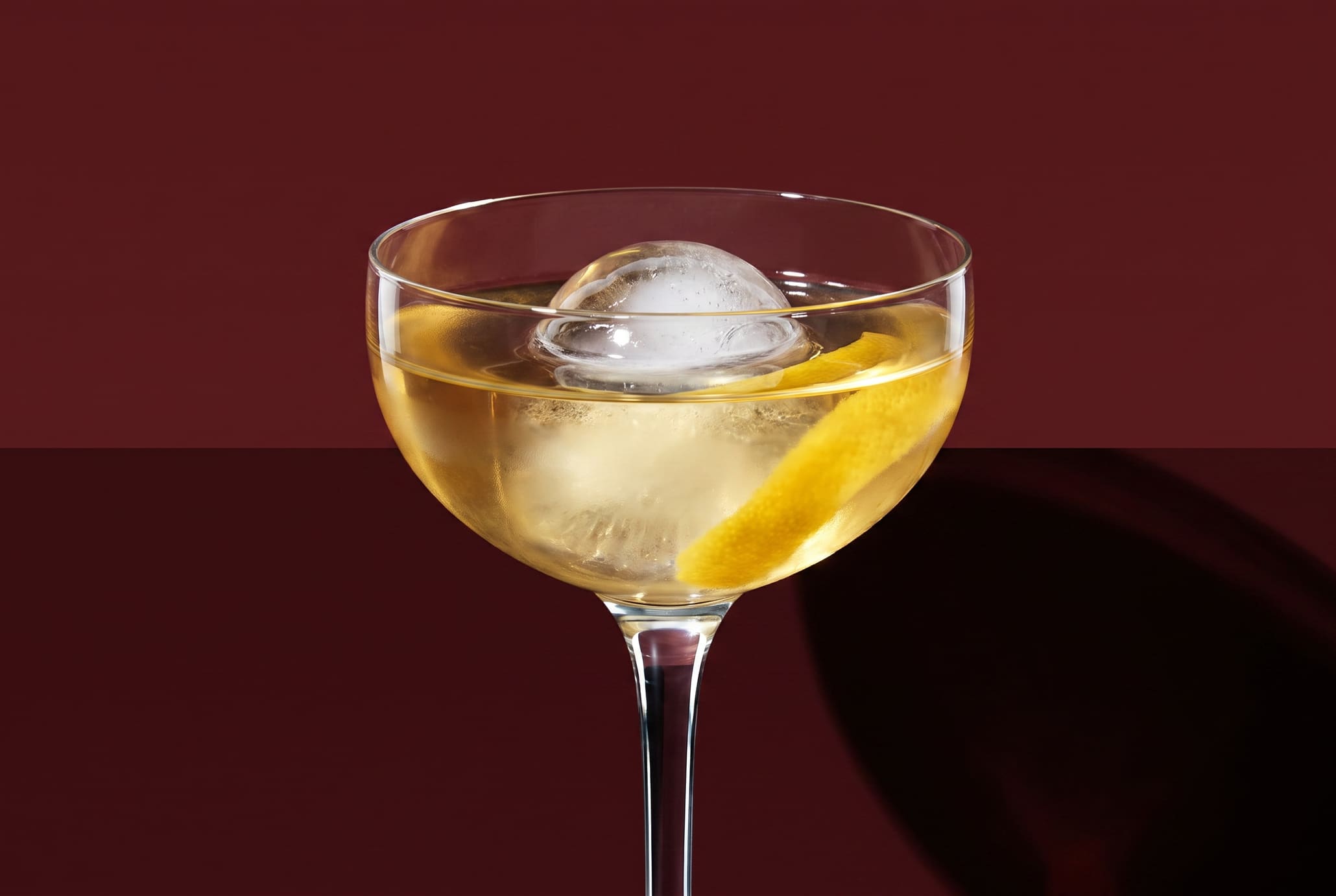 A coupe of pale yellow cocktail with a single ice sphere, lit hard against a deep oxblood backdrop.