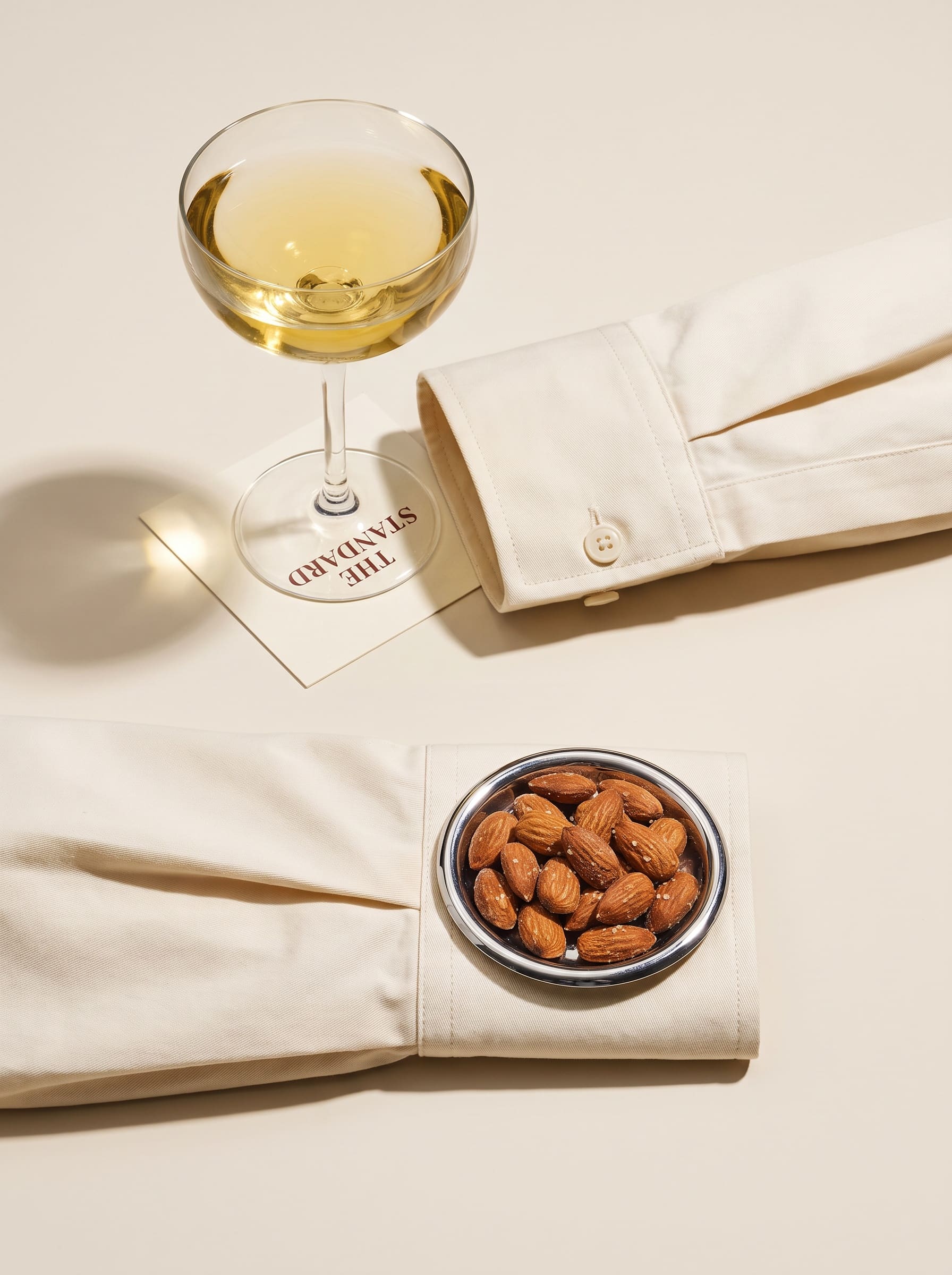 Two crisp shirt cuffs against a flat cream ground, one propping a pale yellow coupe, one supporting a chrome dish of almonds.